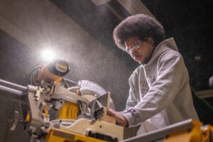 A man wearing a hoodie and safety glasses while working with a table saw.