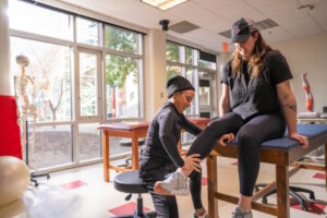 A woman seated on a table with another woman kneeling in front of her to help stretch her leg