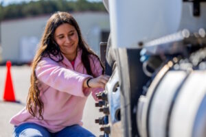 A woman kneeling beside the tire of a tractor trailer.