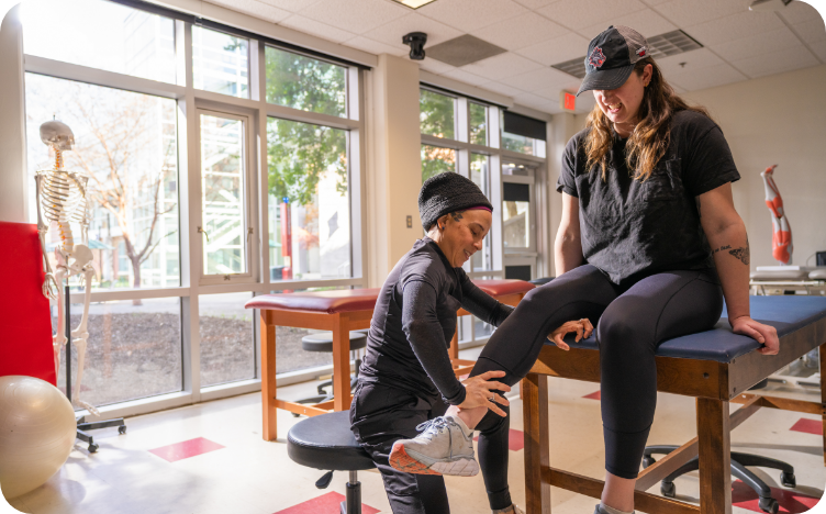 A woman seated on a table with another woman kneeling in front of her to help stretch her leg