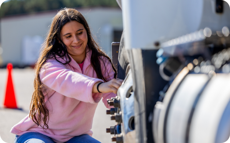 A woman kneeling beside the tire of a tractor trailer.