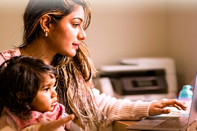 A student working at a laptop with her young daughter beside her