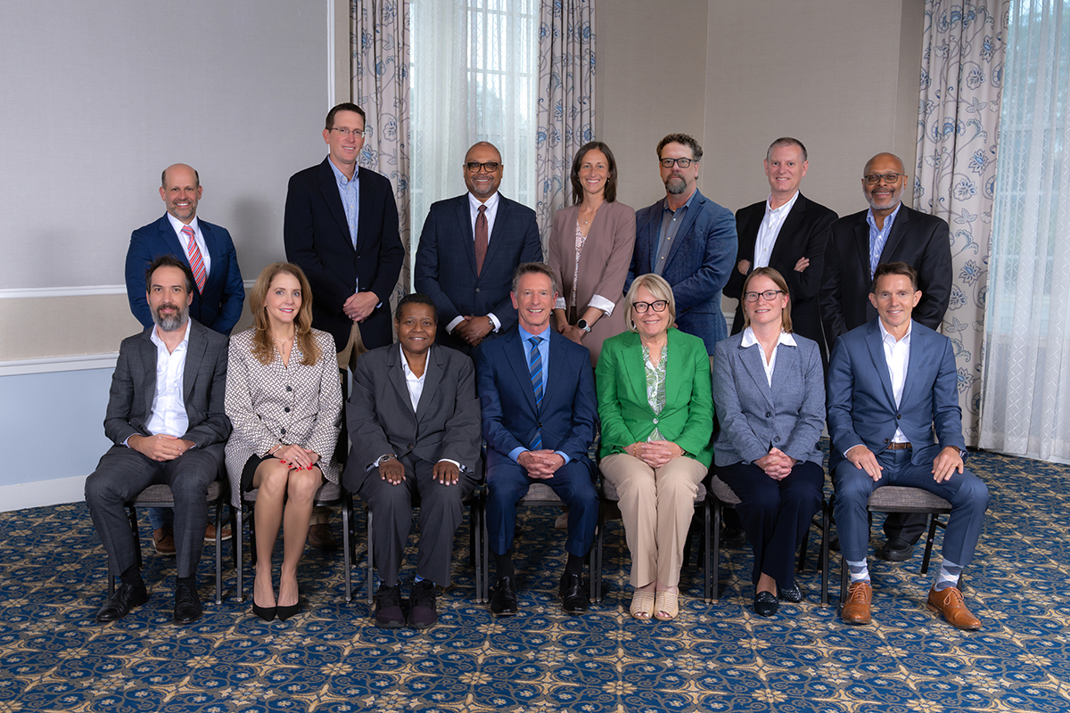 The 14 members of the VCCS Chancellor's Cabinet standing and seated together in business attire in a meeting room.